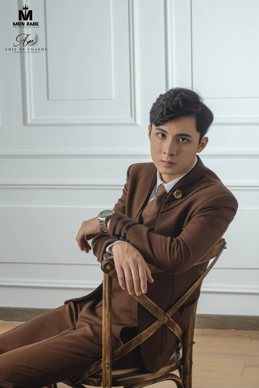 Suit nâu ngồi ghế gỗ bắt chéo, chống tay nhìn thẳng ống kính