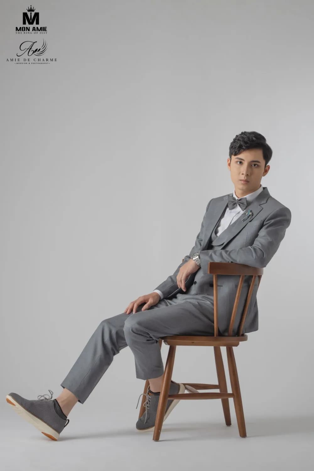 Suit ba mảnh xám, nơ xám, ngồi nghiêng trên ghế gỗ tựa nan