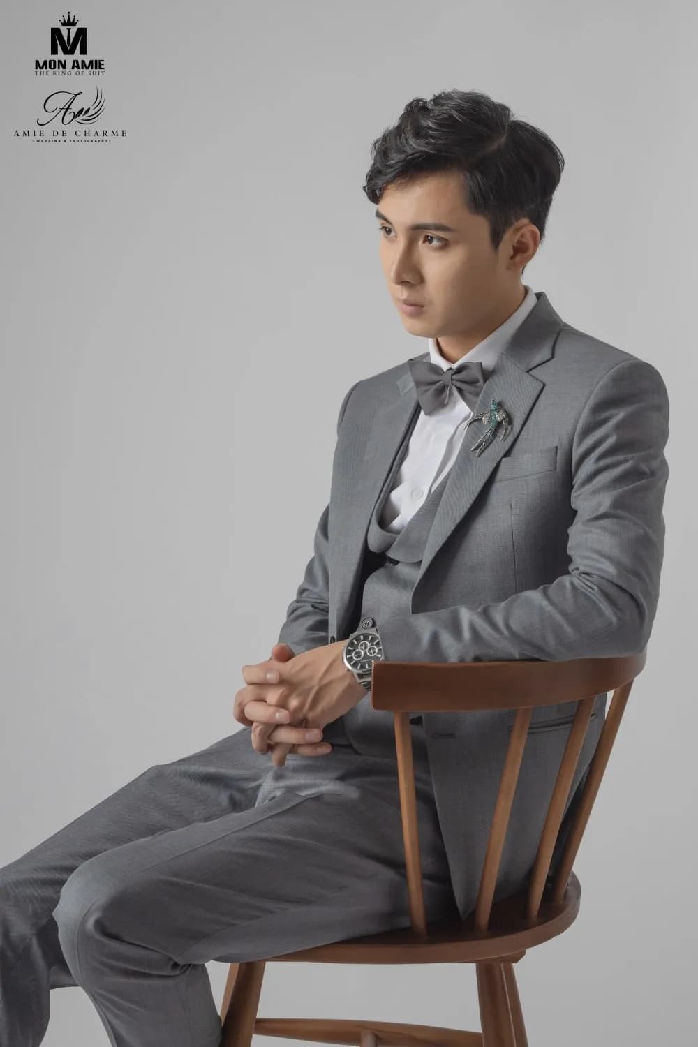 Suit ba mảnh xám, nơ, chắp tay ngồi nghiêng trên ghế gỗ, nhìn xa