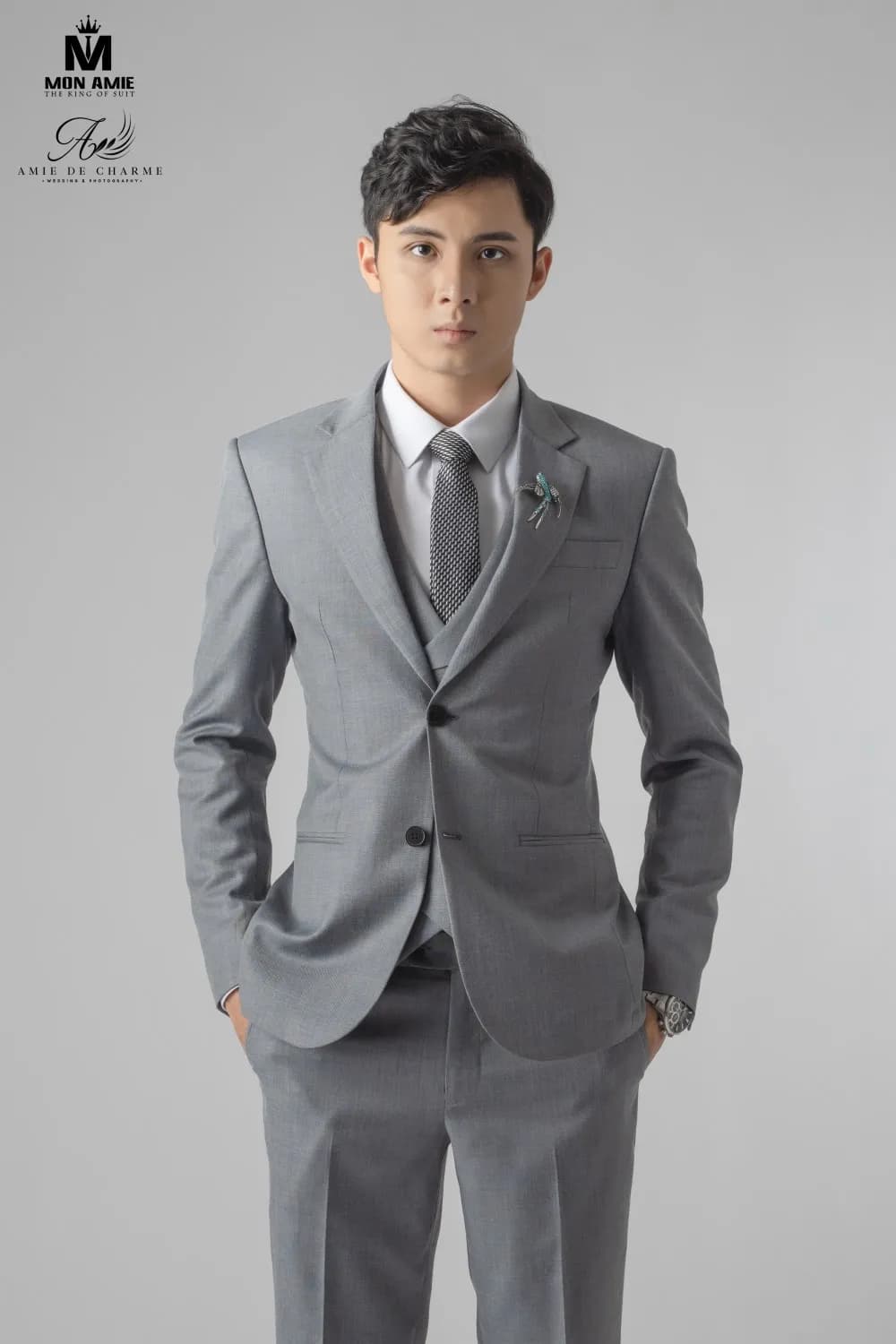 Suit xám ba mảnh, cài hoa ve áo, tay đút túi, nhìn thẳng