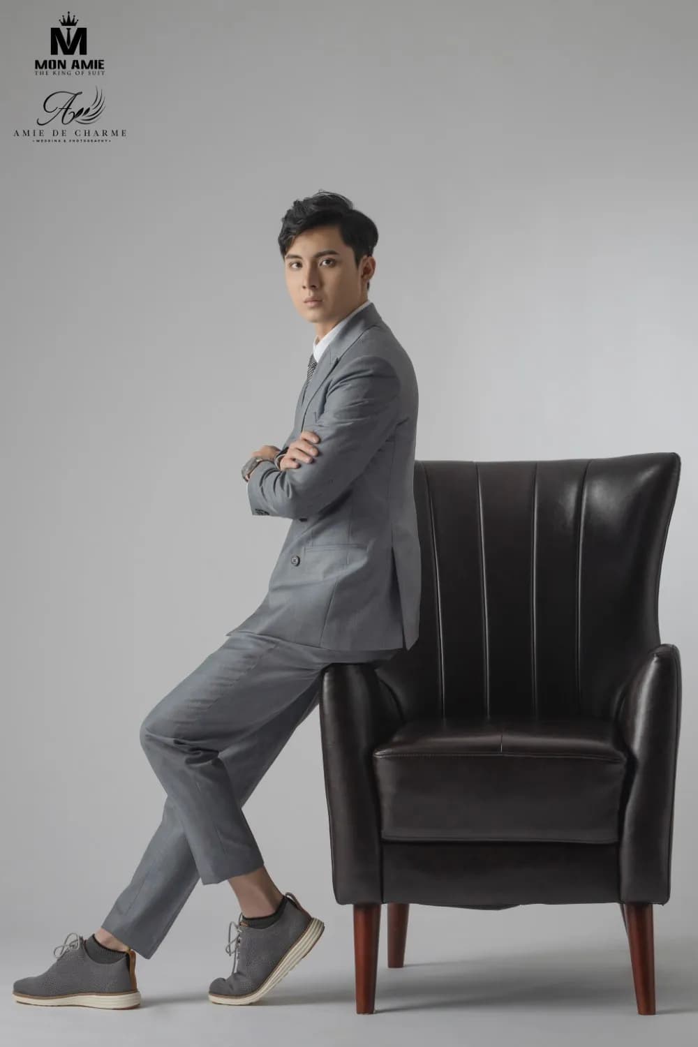 Suit xám, khoanh tay tựa lưng ghế bành da, dáng đứng nghiêng