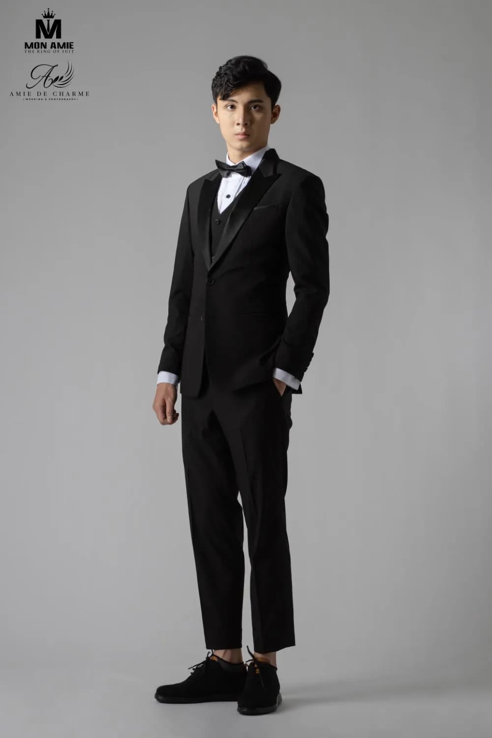 Tuxedo đen thắt nơ, đứng toàn thân, tay đút túi, studio xám