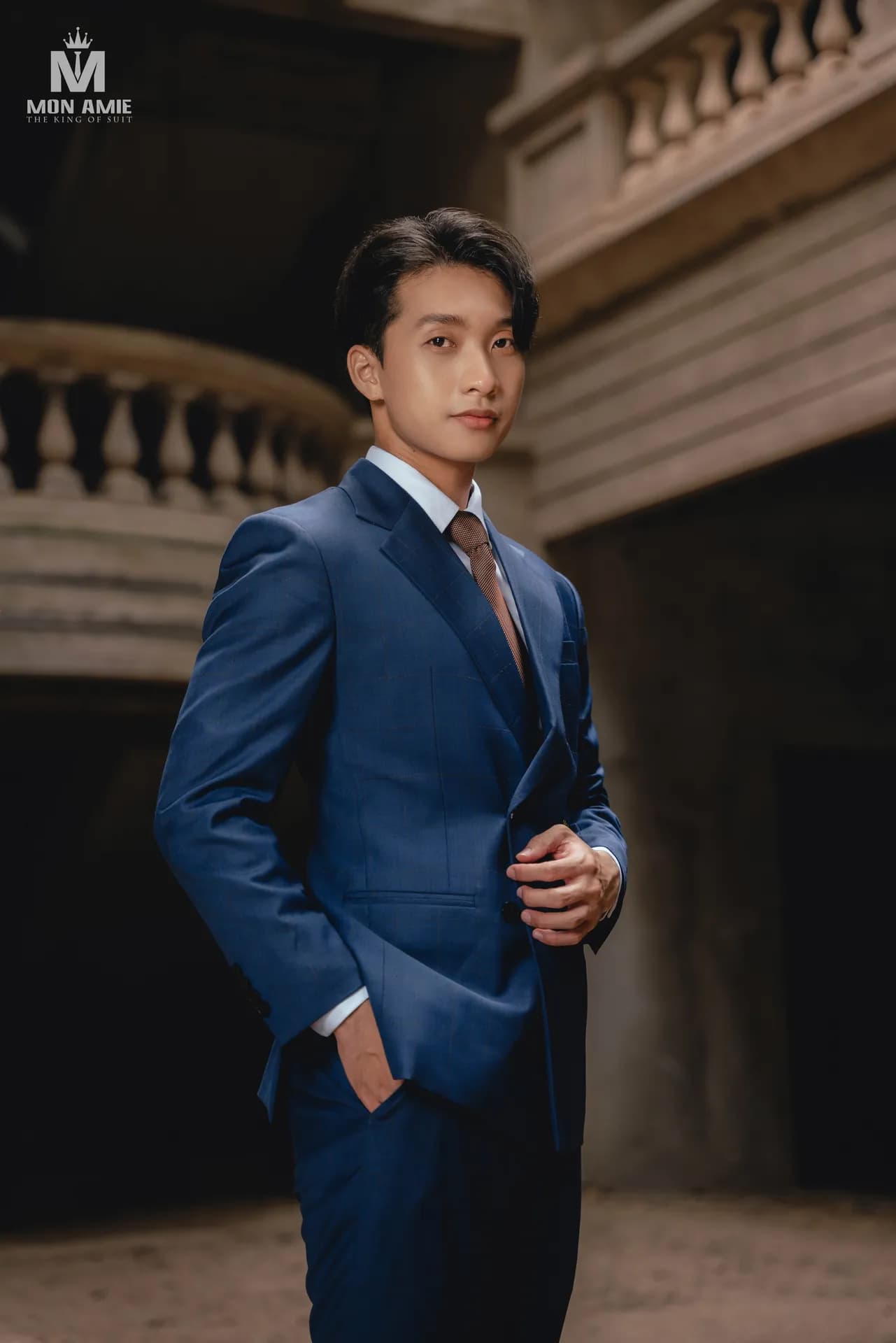 Suit xanh sapphire, cà vạt nâu, tay đút túi trong không gian cổ kính tối