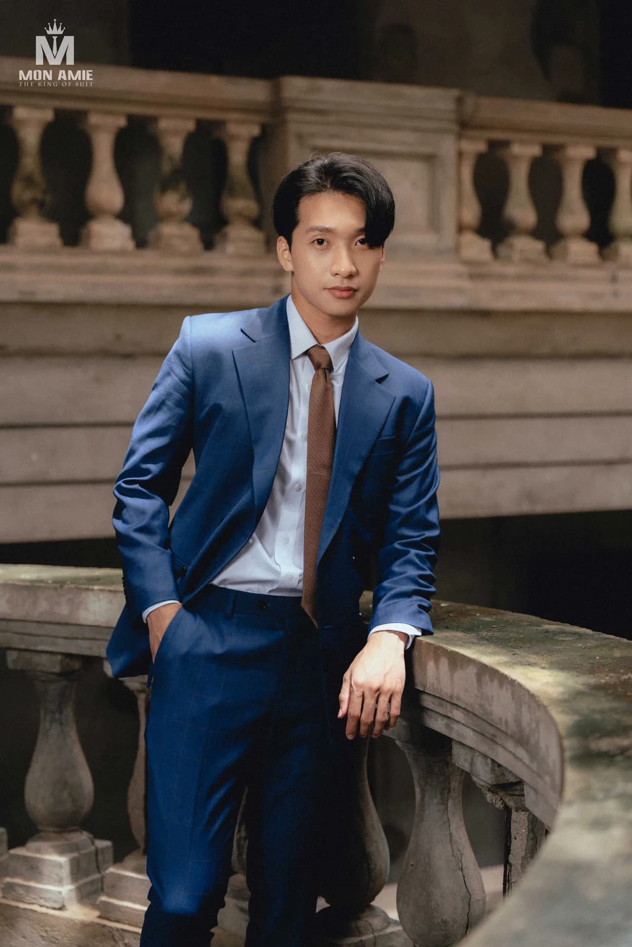Suit xanh sapphire, cà vạt nâu, dựa lan can đá cổ