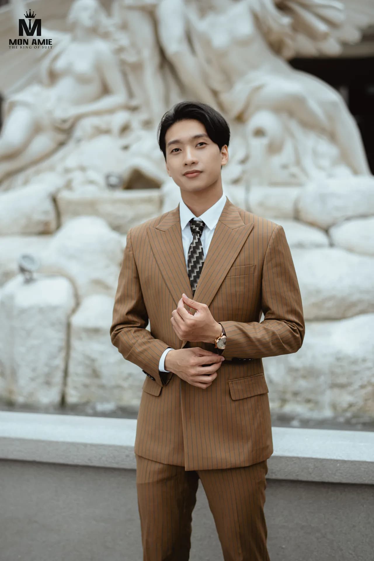 Suit nâu kẻ sọc hai hàng khuy, cà vạt zigzag, đồng hồ vàng hồng, trước phù điêu trắng
