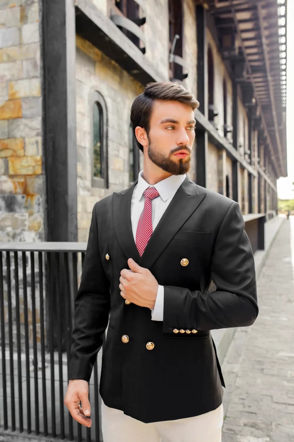 Blazer đen hai hàng khuy vàng, cà vạt sọc đỏ trước tường đá