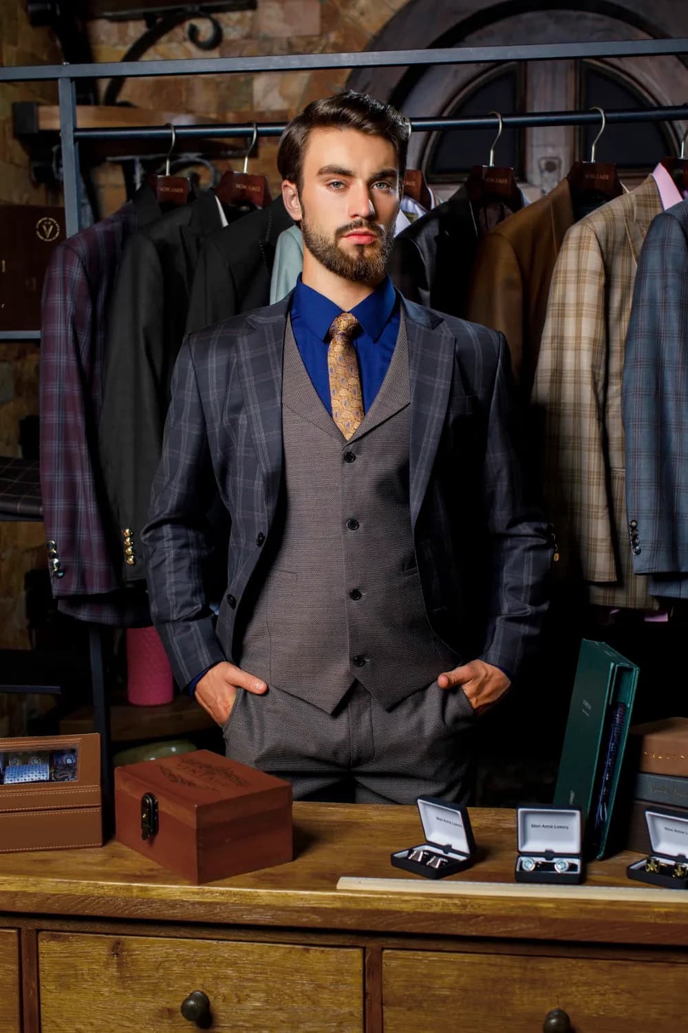Suit ba mảnh kẻ ô xanh đen, gilê xám nâu, tay đút túi trong xưởng may