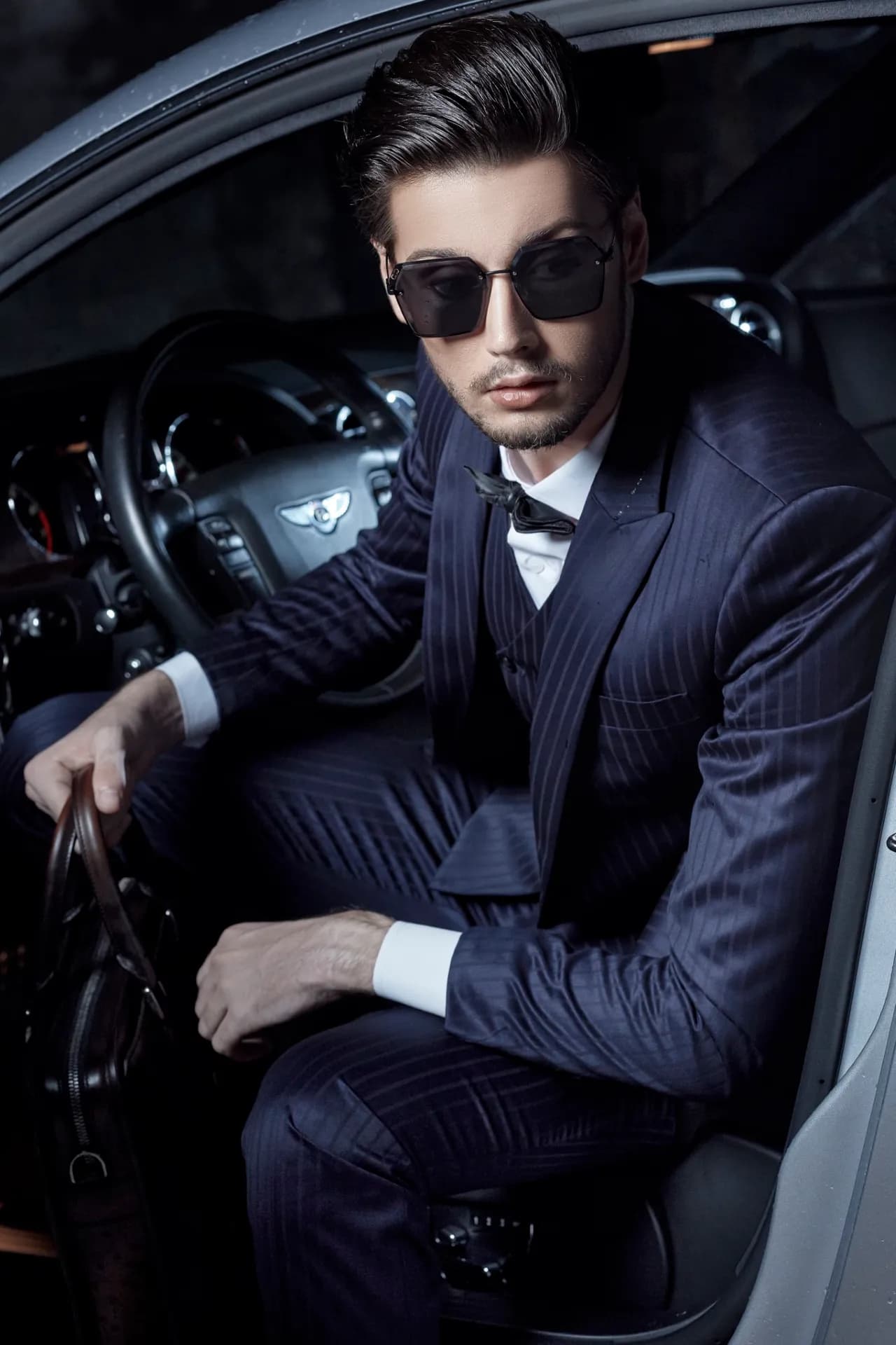 Suit navy kẻ sọc, nơ đen, kính râm, ngồi trong xe sang cầm cặp da