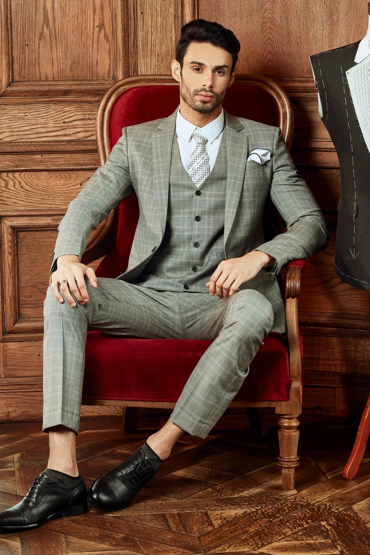 Bộ suit ba mảnh caro xám ngồi trên ghế nhung đỏ, khăn cài túi trắng