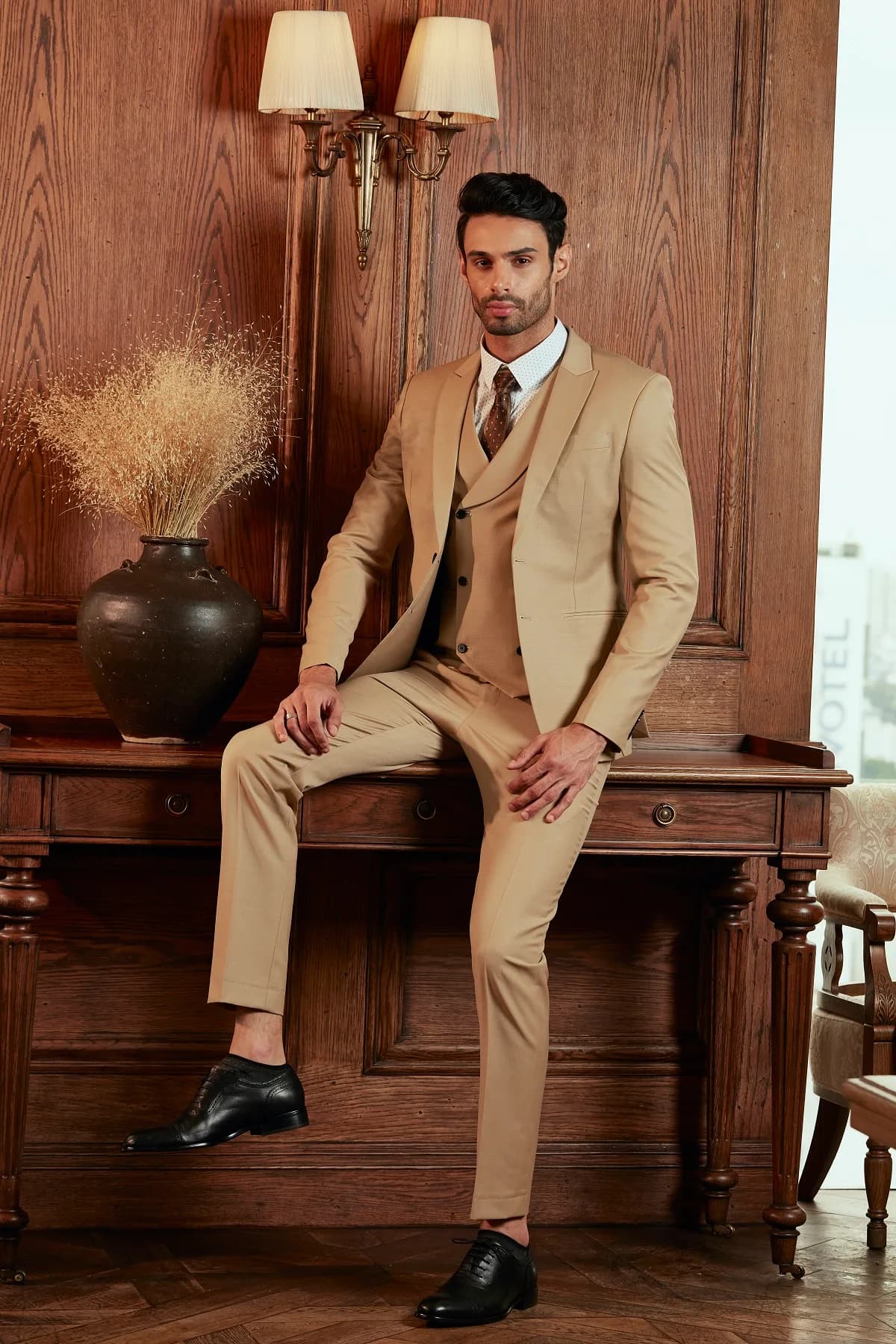 Suit ba mảnh màu be camel, dáng ngồi tựa bàn gỗ cổ điển