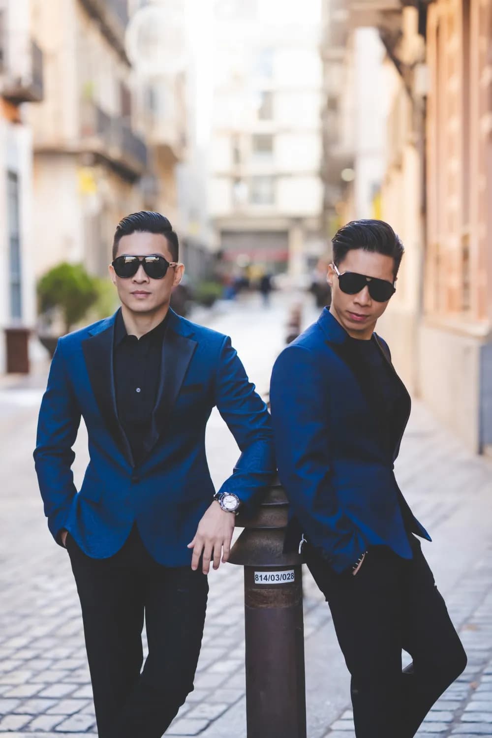 Hai blazer xanh paisley cổ ve đen, tựa trụ sắt trên phố lát đá