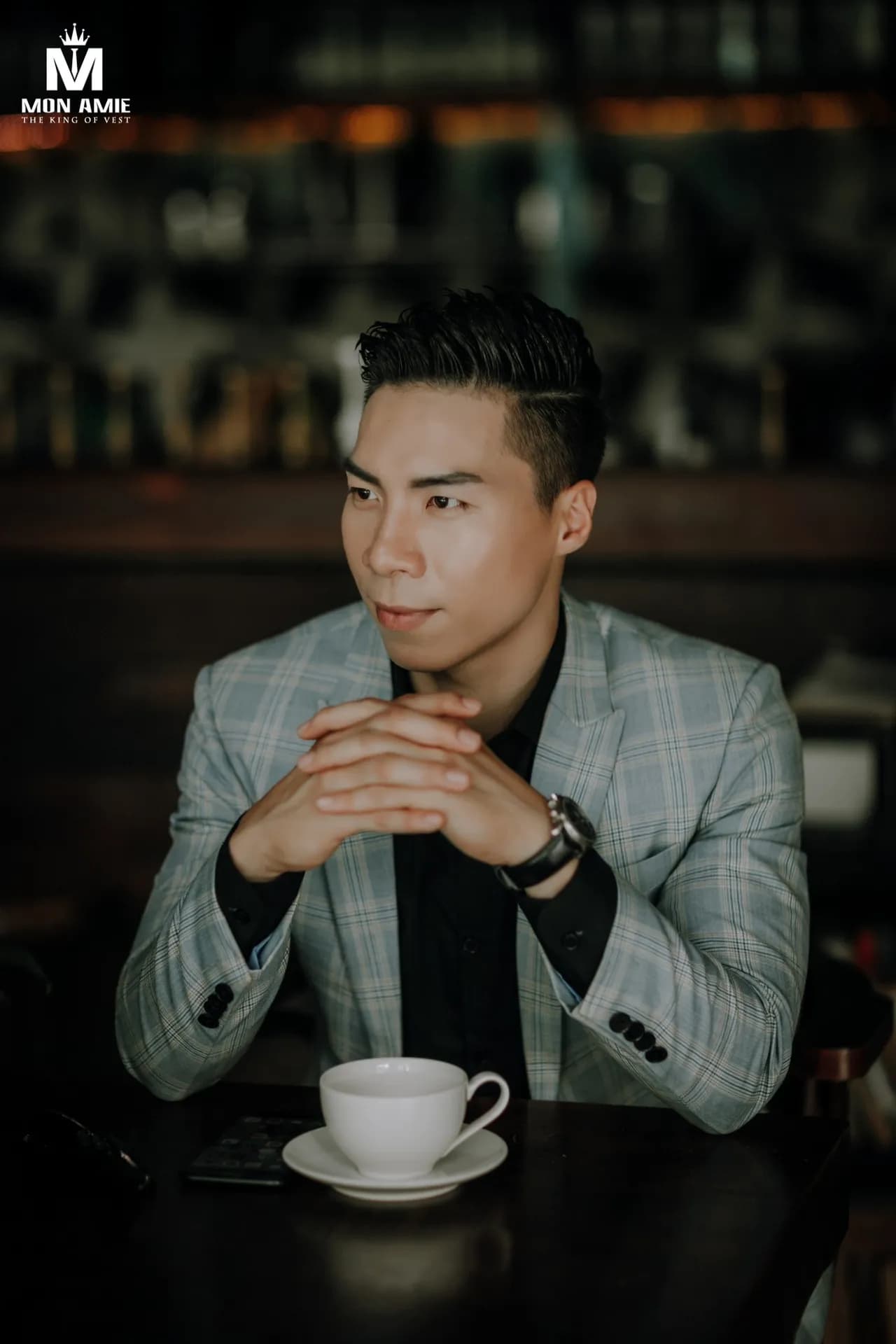 Blazer caro xanh xám, sơ mi đen, chắp tay bên tách cà phê trong quán tối