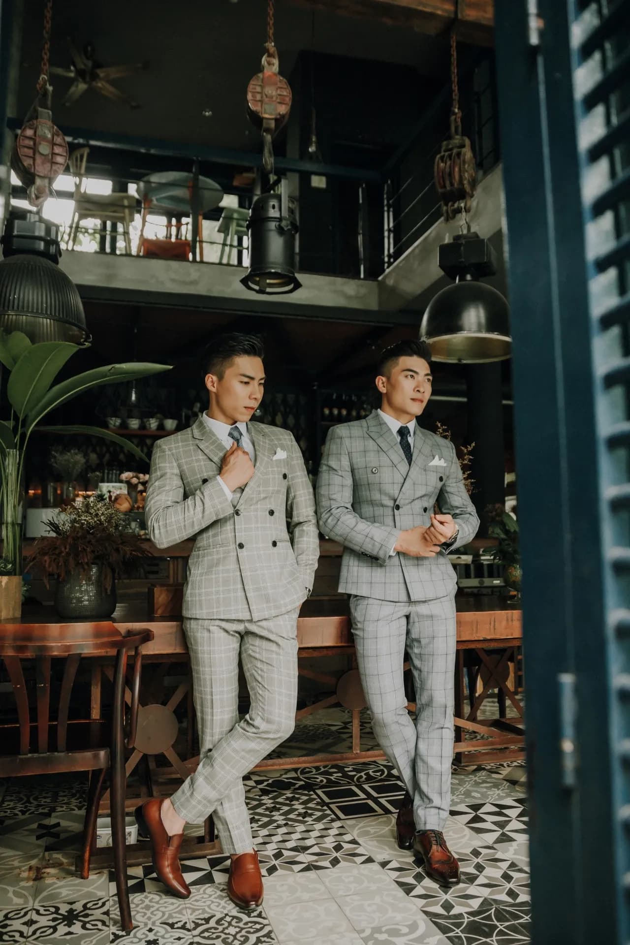 Hai suit caro xám hai hàng khuy, khăn túi trắng, tựa bàn gỗ trên sàn gạch hoa