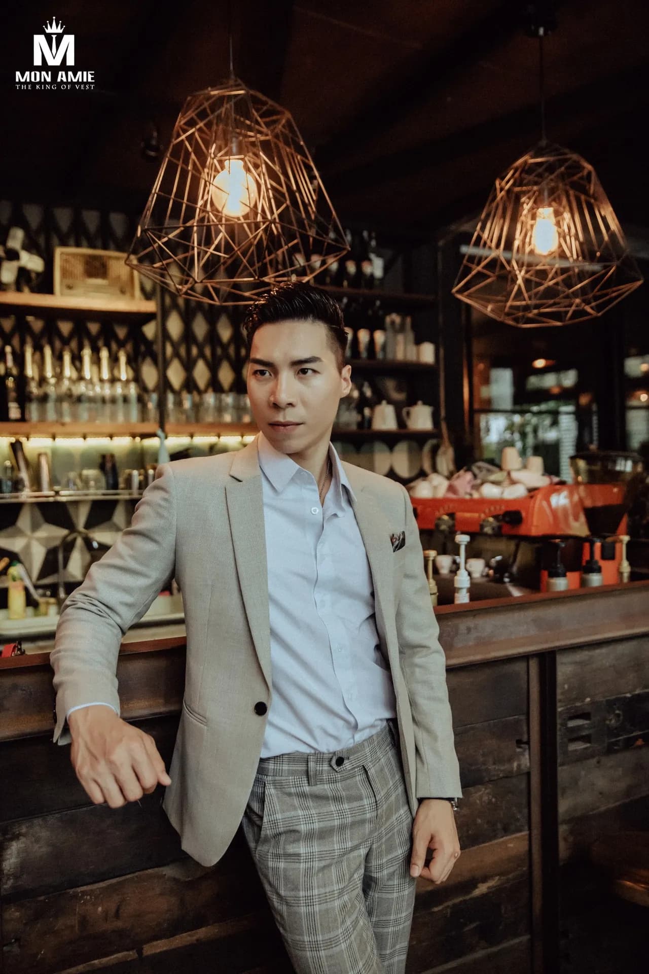 Blazer be, quần caro, sơ mi xanh nhạt, dựa quầy bar dưới đèn lồng