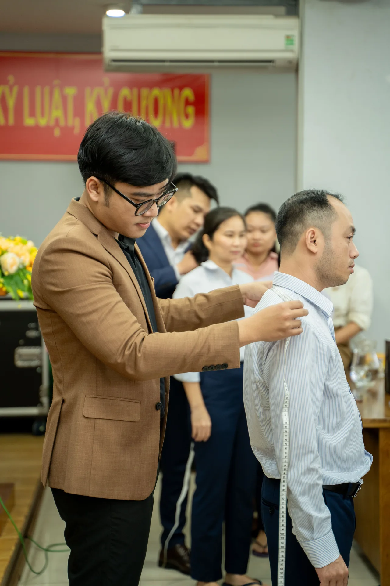 Mon Amie staff member measuring the shoulders of a male employee while colleagues line up behind