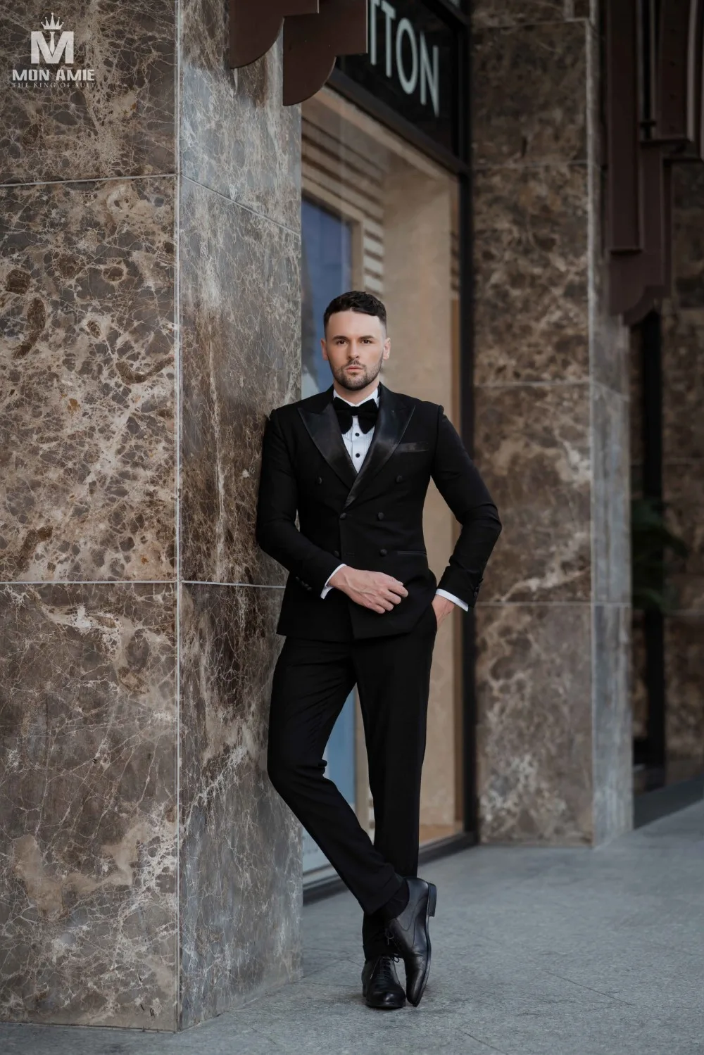 Tuxedo đen tựa cột đá cẩm thạch, giày da bóng và nơ đen, minh họa vest đen mang giày đen bóng cho dịp trang trọng