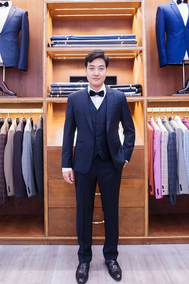 Suit ba mảnh xanh navy đậm, nơ đen, đứng trước kệ vải gỗ, minh họa vest chú rể may đo cho ngày cưới