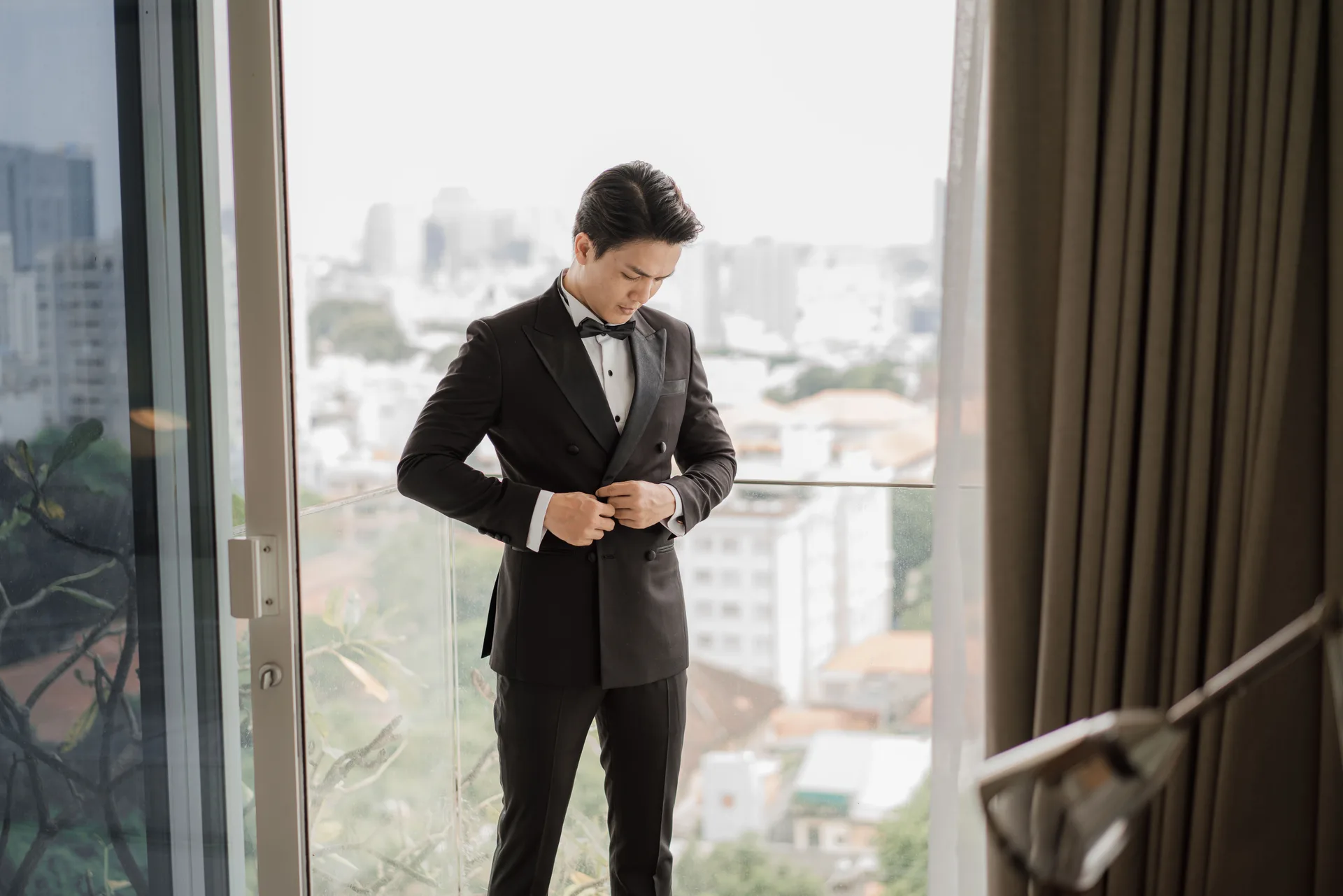 A gentleman adjusting the buttons of a double-breasted tuxedo jacket on a balcony overlooking the city