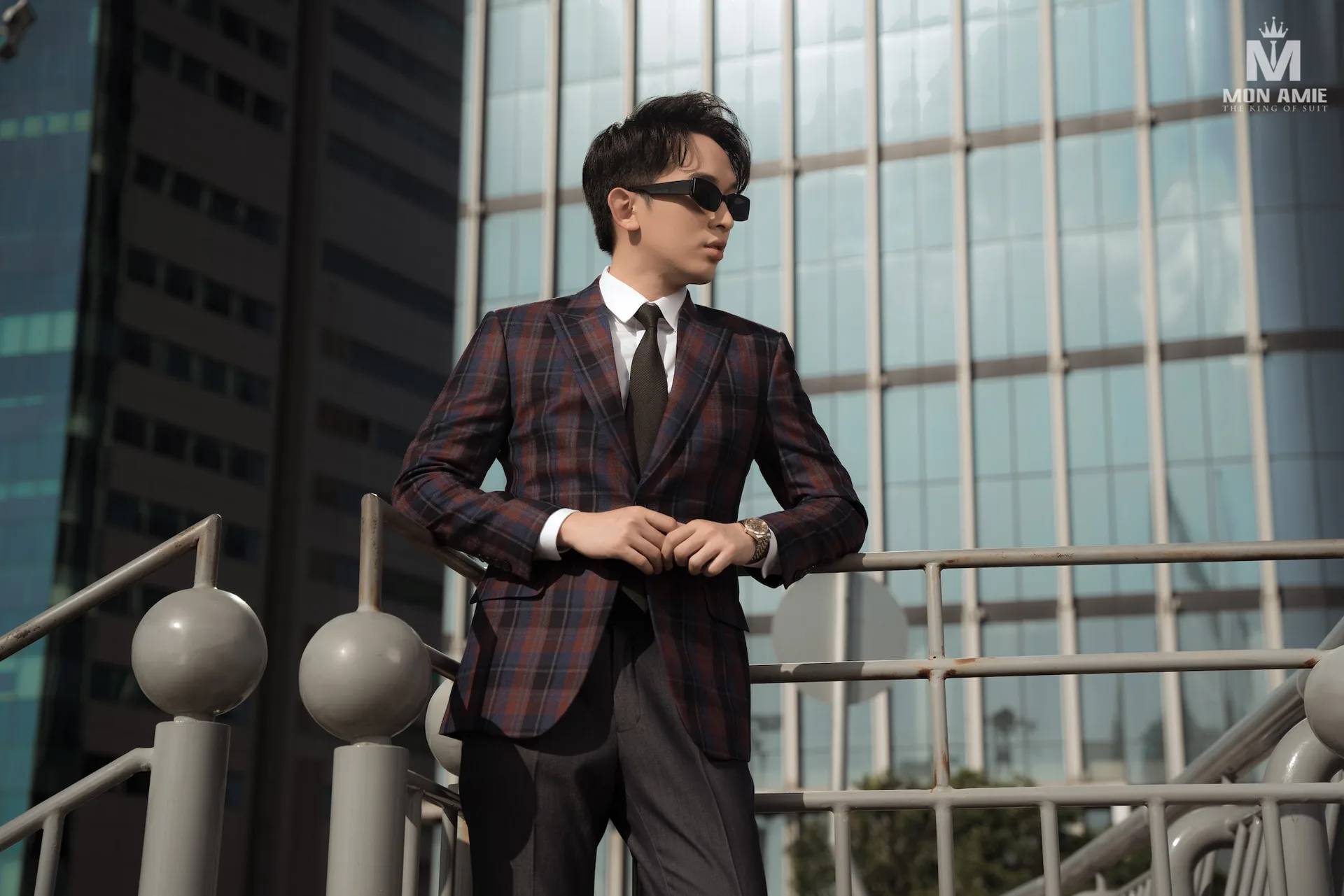 Blazer caro tím nâu phối quần xám, dáng tựa lan can giữa các tòa nhà kính
