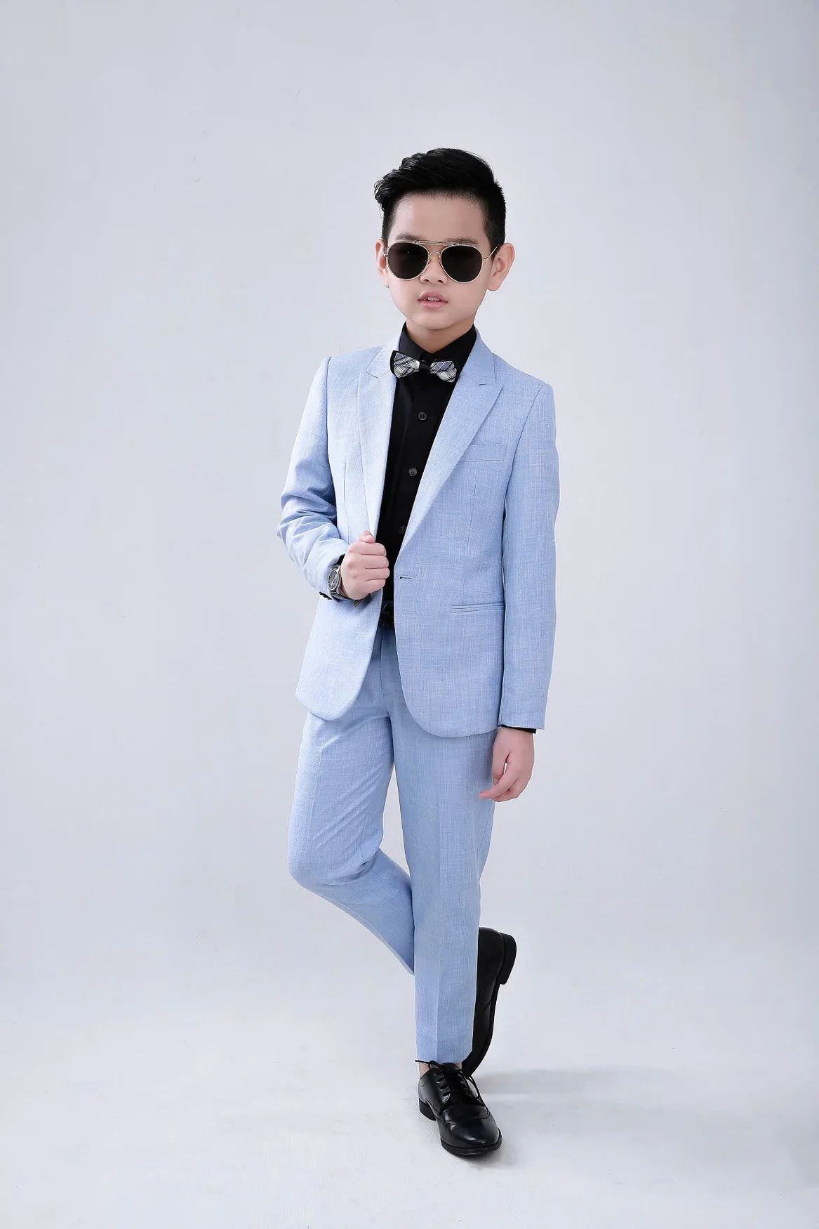 Boy in a pastel blue suit, black shirt and check bow tie against a white backdrop