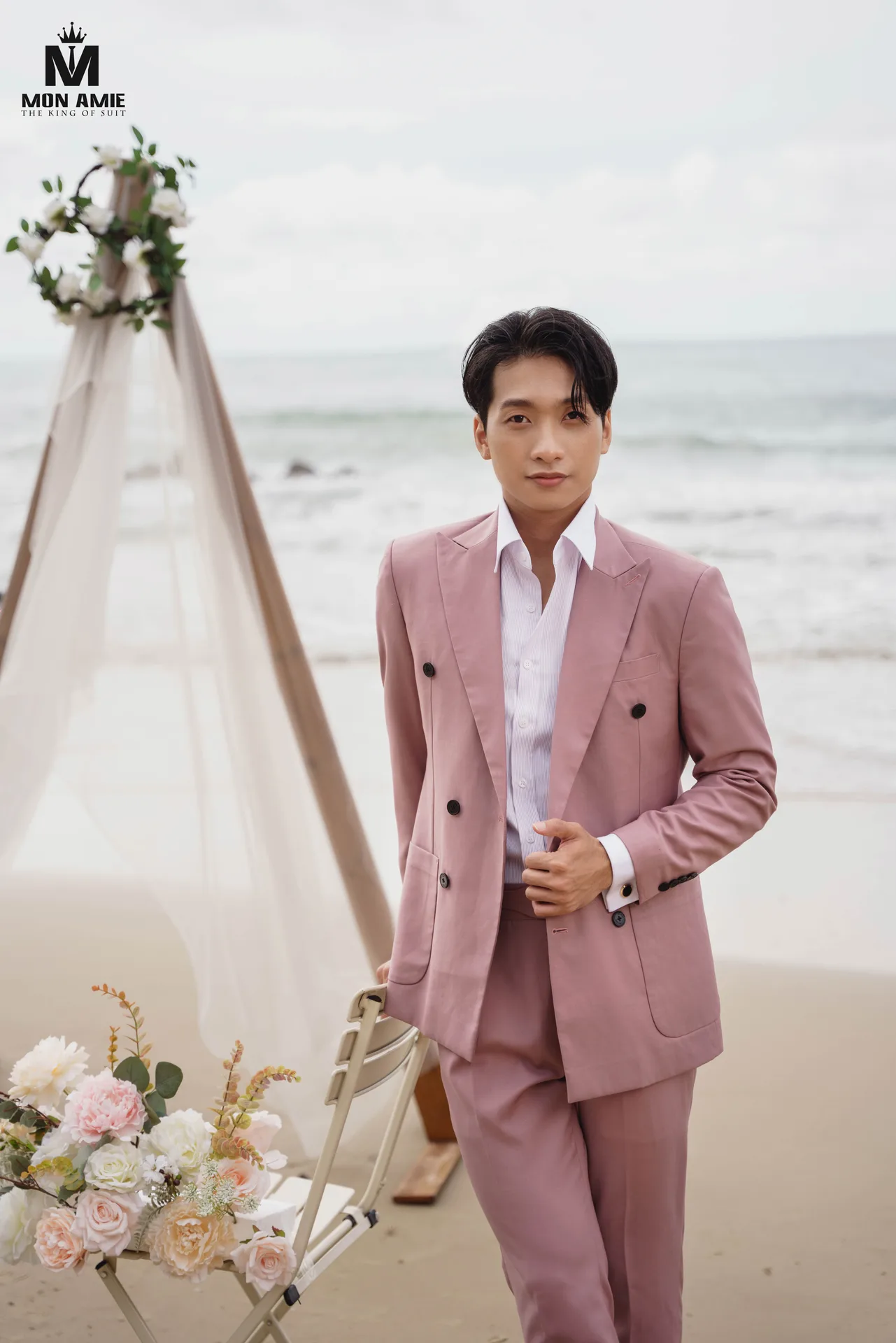Earthy pink double-breasted suit on a beach beside a voile flower arch, an earthy pink suit for men at an outdoor party